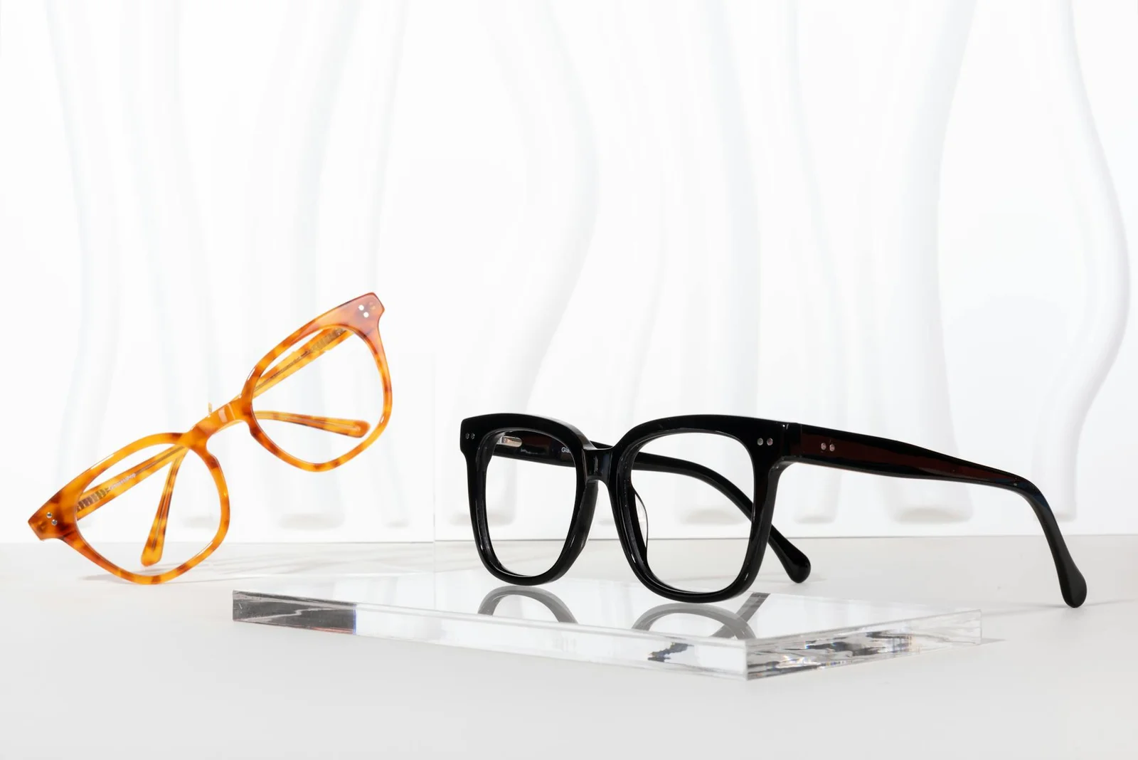 Elegant glass frames in black and tortoise colors showcased on a clear acrylic stand against a wavy backdrop.